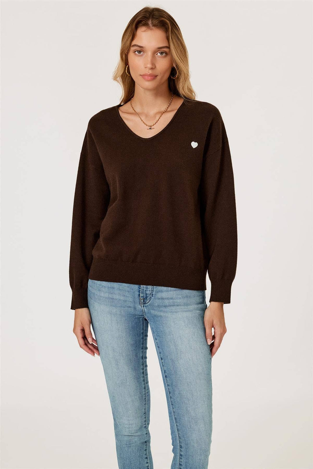 Woman wearing a brown sweater with a heart emblem and light blue jeans on a white background