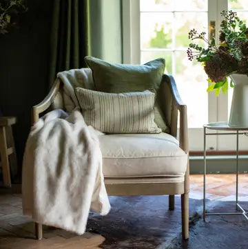 A light beige upholstered armchair with wooden arms in a styled room, featuring a cream throw blanket draped over the arm, a striped accent pillow, and a solid green pillow. A small side table with flowers is visible to the right.