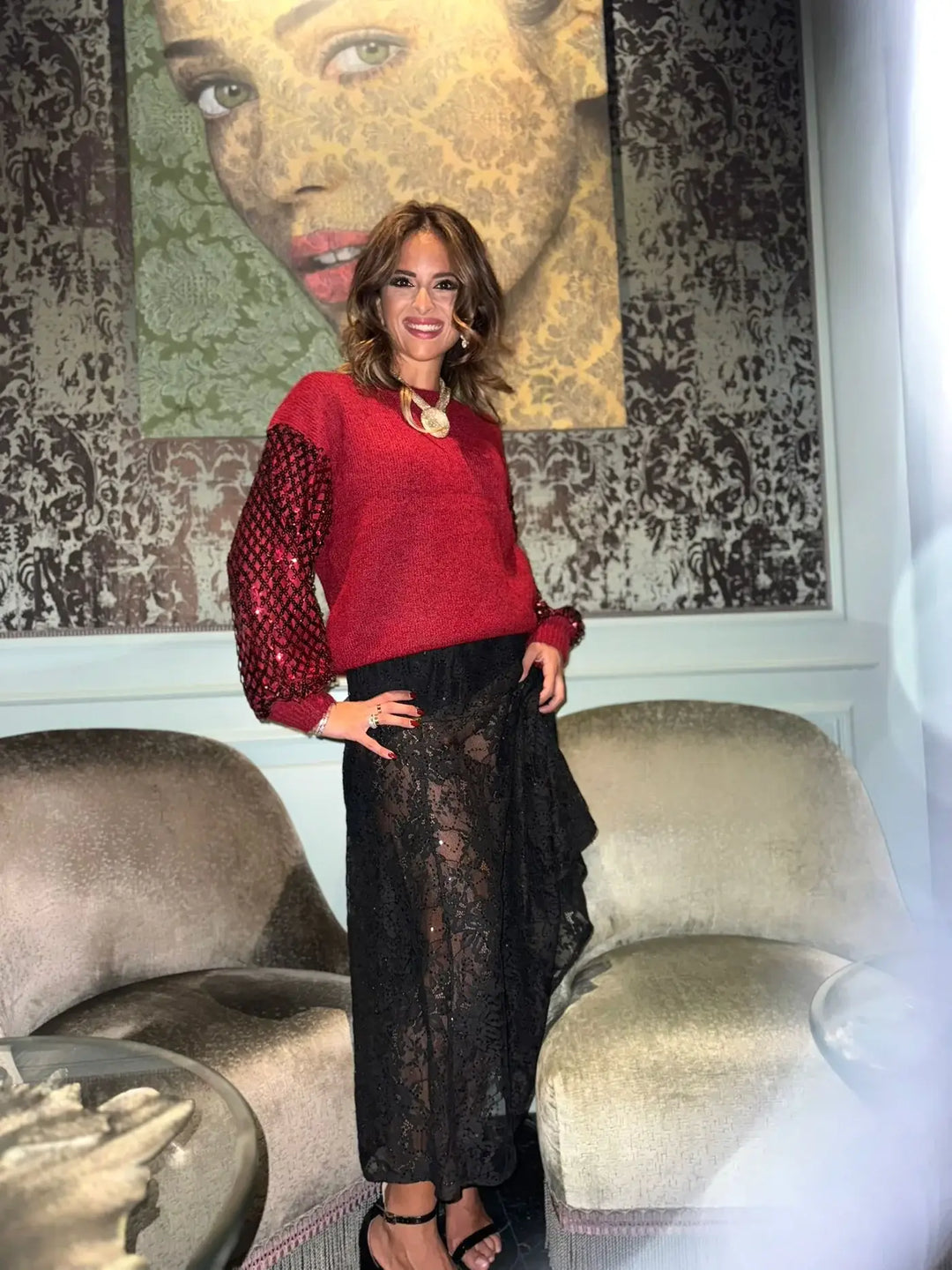 Woman in a red sweater and black lace skirt standing in a room with a textured wall and sofa.