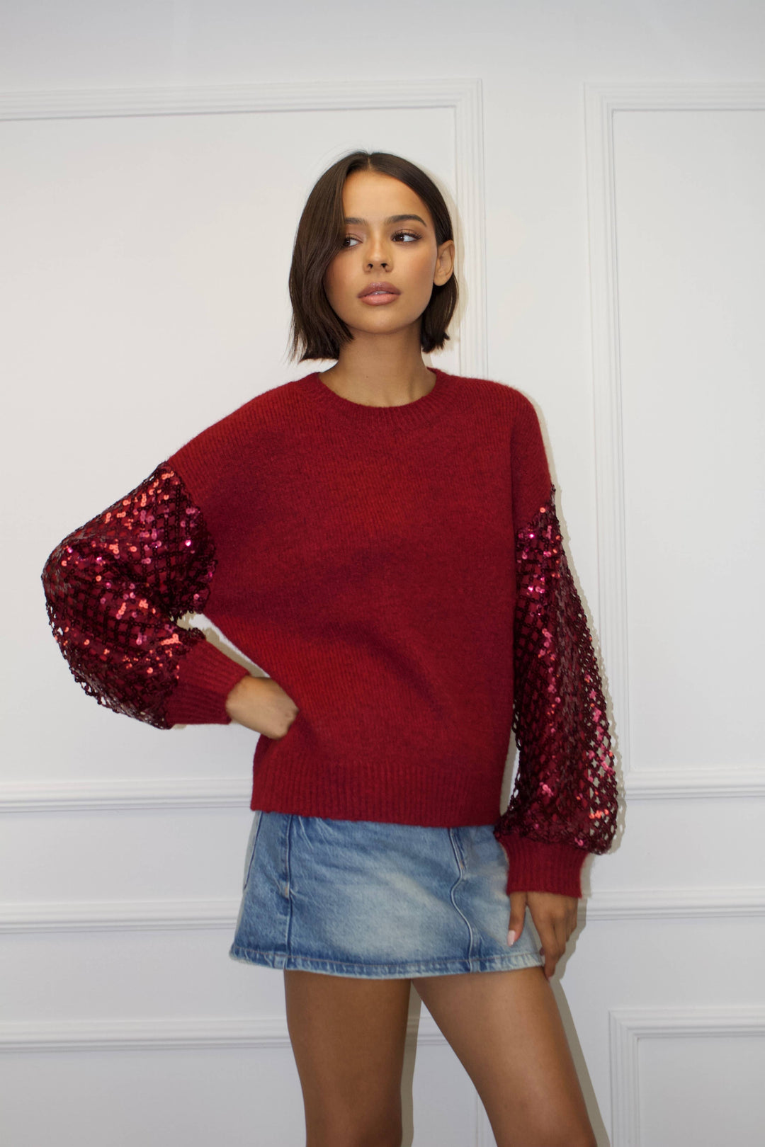 Woman wearing a red sweater with sequin sleeves and a denim skirt against a white background