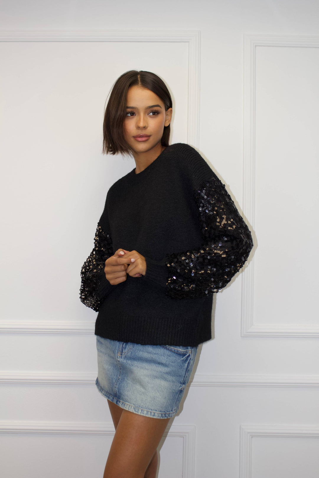 Woman wearing a black sweater with sequin sleeves and a denim skirt against a white paneled wall.