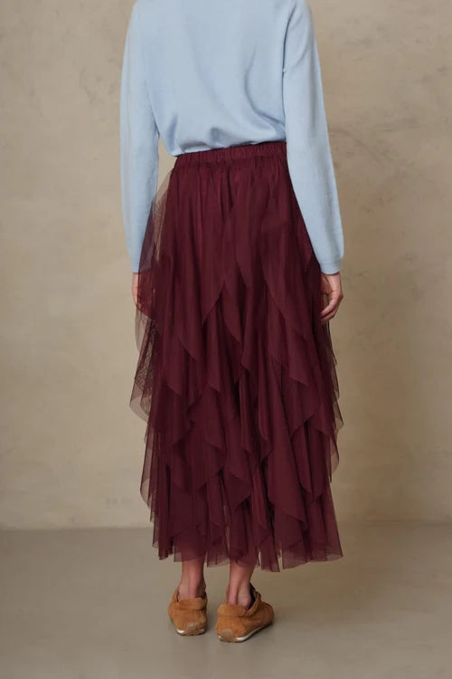 A person shown from the back wearing a long, tiered tulle midi skirt in deep burgundy, paired with a light blue long-sleeve top and tan shoes, against a neutral textured beige studio backdrop.