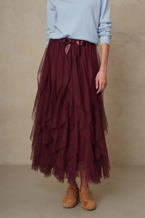 A model wearing a long, tiered tulle midi skirt in deep burgundy, paired with a light blue long-sleeve top and brown suede boots, standing against a textured beige wall.