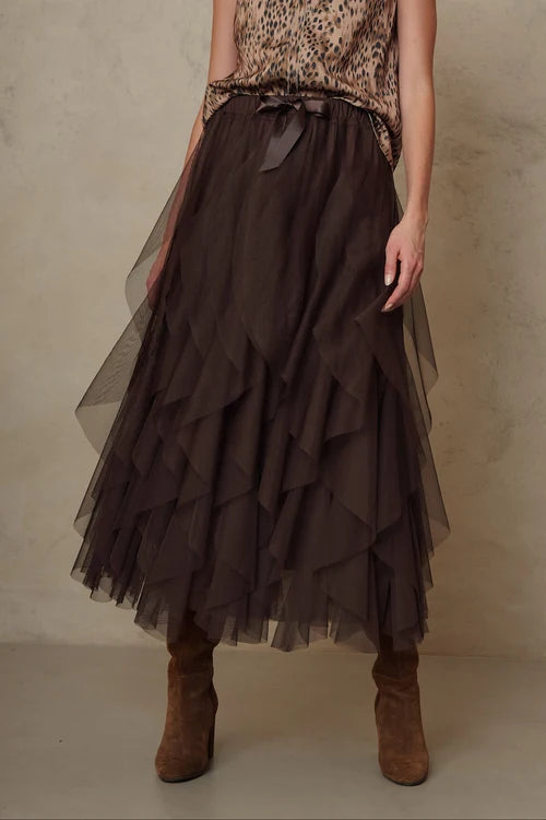 A model wearing a dark brown tiered tulle midi skirt with a matching fabric tie belt at the waist, paired with a leopard-print top and brown suede boots, set against a textured beige studio backdrop.
