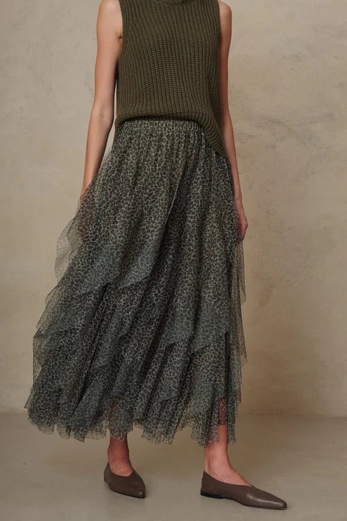 A model wearing a sleeveless olive green ribbed knit top and a long, tiered dark gray-green tulle skirt with a subtle floral pattern, paired with brown leather flats, set against a textured beige studio backdrop.