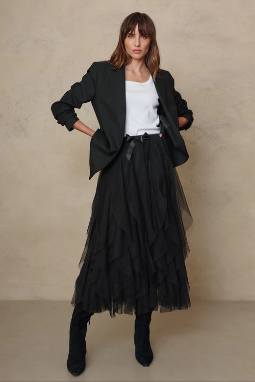 A woman models a black long-sleeve blazer, a white ribbed top, a long black tiered tulle midi skirt with a black ribbon tie, and black knee-high boots, set against a textured beige studio backdrop.