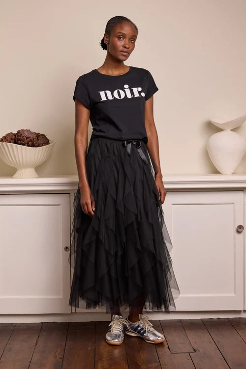 Woman wearing a black t-shirt with 'noir' text and a black tulle skirt in a room with white cabinets and decorative items.