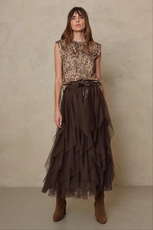 A woman stands against a textured beige wall wearing a sleeveless leopard-print top and a long dark brown ruffled tulle skirt with a matching belt, paired with brown ankle boots.