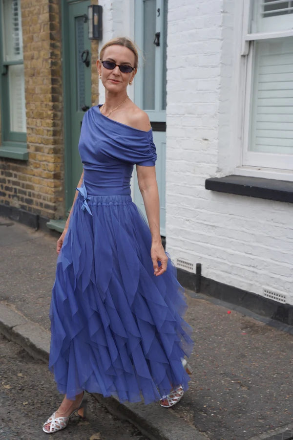 A woman wearing a royal blue off-the-shoulder maxi dress with a ruffled tiered skirt, standing on a street in front of a white brick house.