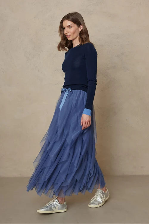 A woman modeling a navy blue long-sleeve knit top and a layered blue tulle midi skirt with a matching blue ribbon tie, paired with metallic silver sneakers, against a textured beige studio backdrop.