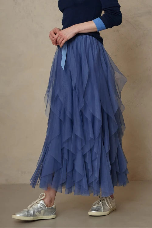 A person wearing a long, tiered tulle midi skirt in dusty blue, paired with a navy top and metallic silver sneakers, standing against a neutral beige textured wall.