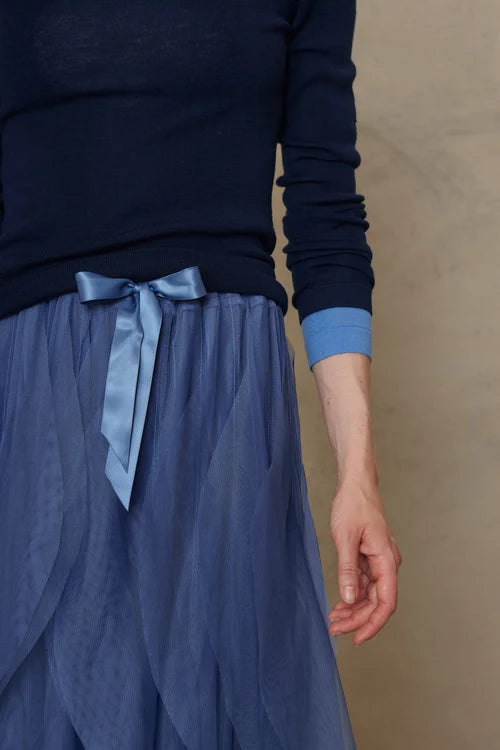 A person wearing a dark navy long-sleeve top and a flowing dusty blue skirt with a matching blue satin bow at the waist, standing against a neutral beige wall.
