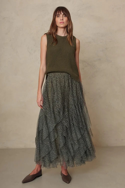 A woman stands against a textured beige wall wearing a sleeveless olive green ribbed knit top and a long dark gray patterned tulle skirt with ruffles, paired with brown pointed flats.