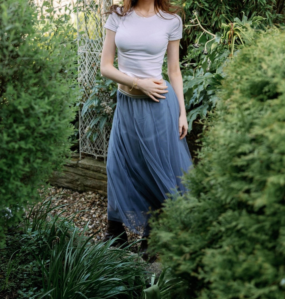 Woman in a garden wearing a light gray top and blue skirt.