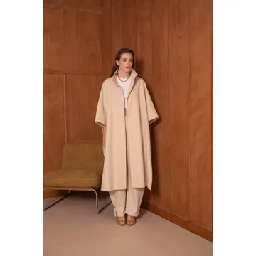 Woman wearing a beige coat and pants standing against a wooden paneled wall.