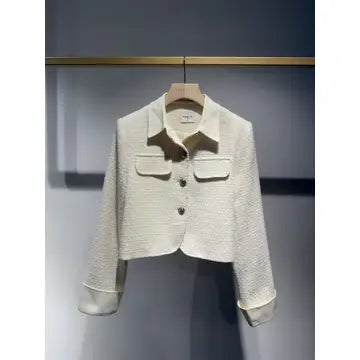 A cropped, long-sleeve textured jacket in off-white/cream, hanging on a wooden hanger against a dark gray wall with a gold horizontal bar above.