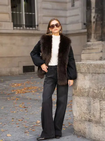 A woman stands outdoors on a stone street corner wearing a black puffer-style jacket with a dark brown fur collar, a white ribbed top, black wide-leg pants, and sunglasses.