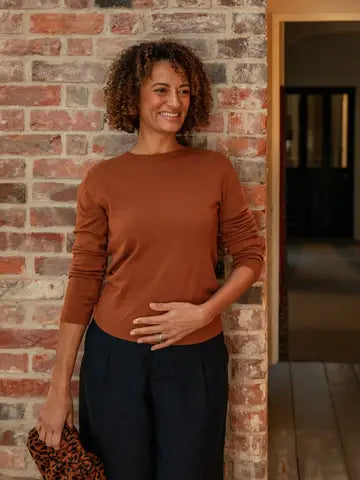 Woman standing against a brick wall wearing a brown sweater and dark pants.