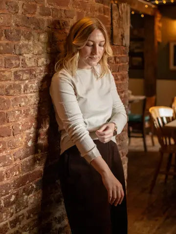 A woman with blonde hair leans against a rustic red brick wall in a warmly lit indoor setting, wearing a cream-colored long-sleeve knit top and dark brown trousers.