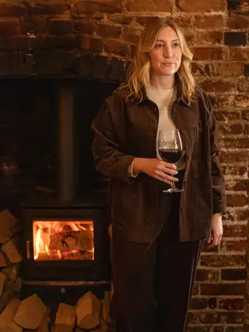 A woman stands in front of a brick fireplace holding a glass of red wine, wearing a dark brown corduroy overshirt over a cream turtleneck and dark trousers.