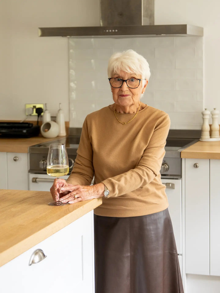 An older woman with short white hair and glasses stands in a modern kitchen, leaning on a wooden countertop and holding a glass of white wine. She is wearing a tan long-sleeve top and a dark brown skirt.