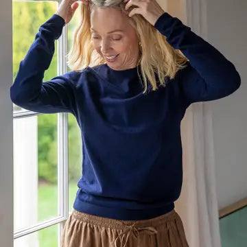 Woman wearing a blue sweater and beige pants indoors with a window in the background