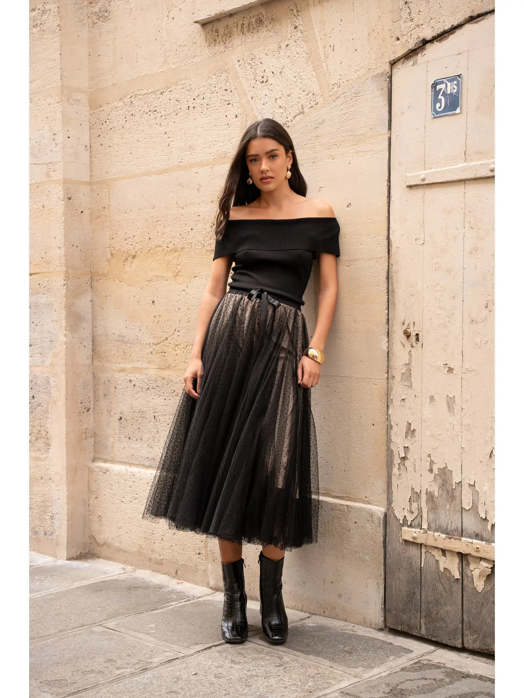 Woman in black off-shoulder top and tulle skirt standing against a textured wall.