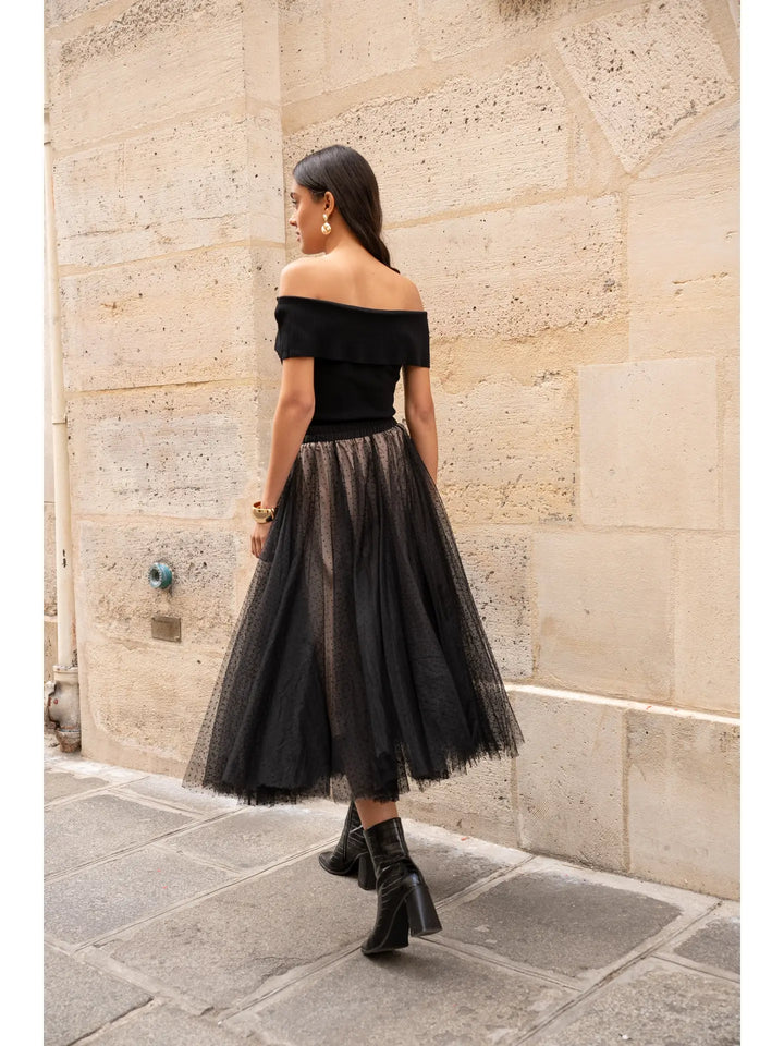 Woman wearing a black off-shoulder top and tulle skirt against a stone wall.