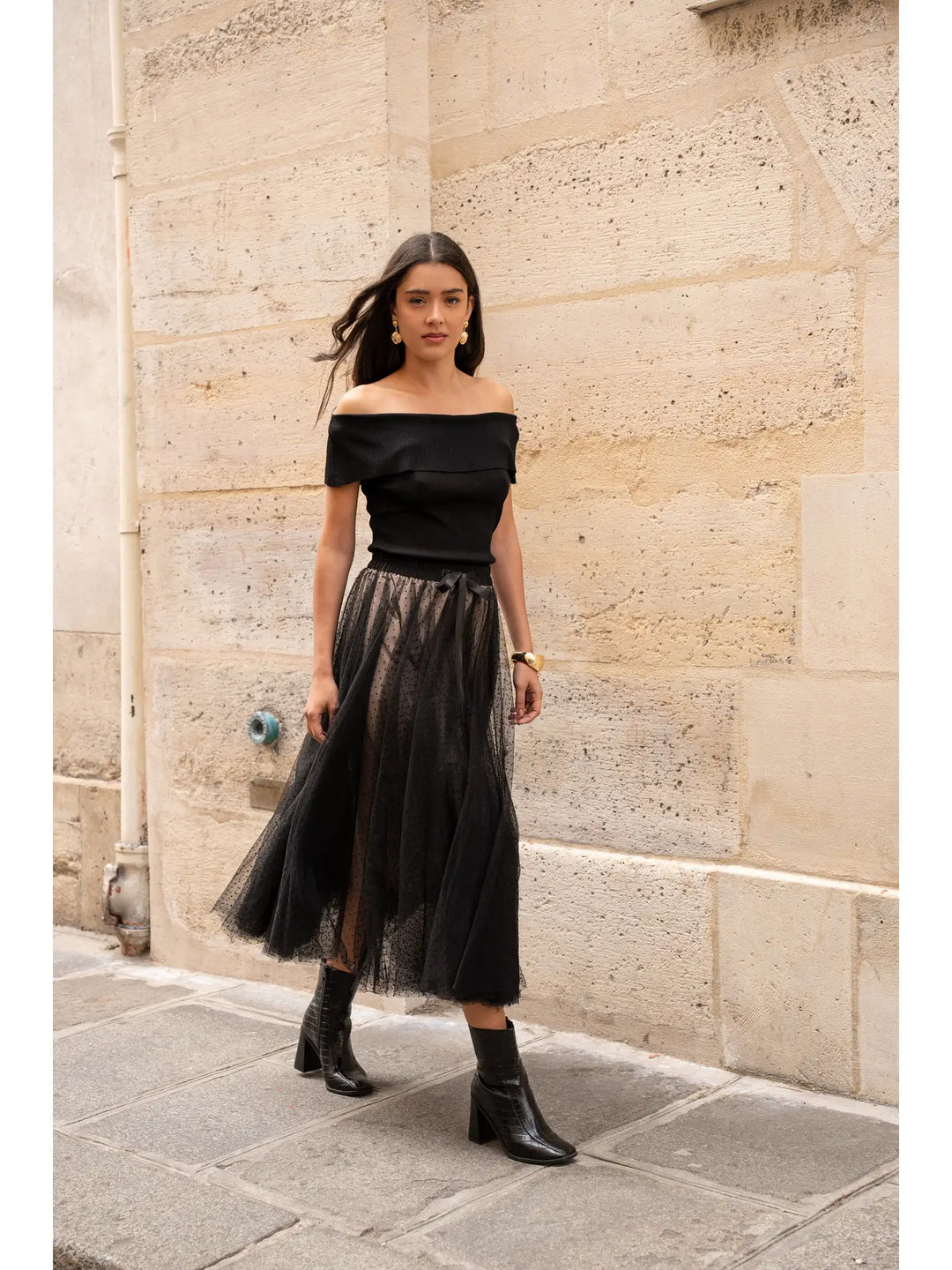 Woman wearing a black off-shoulder top and tulle skirt against a stone wall.