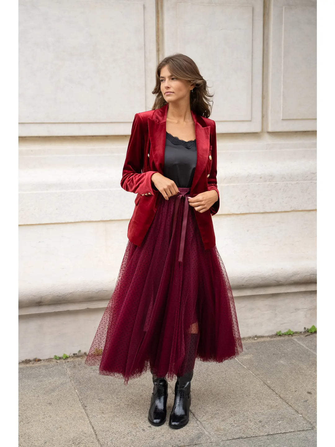 Woman wearing a red blazer and burgundy tulle skirt against a light-colored wall.
