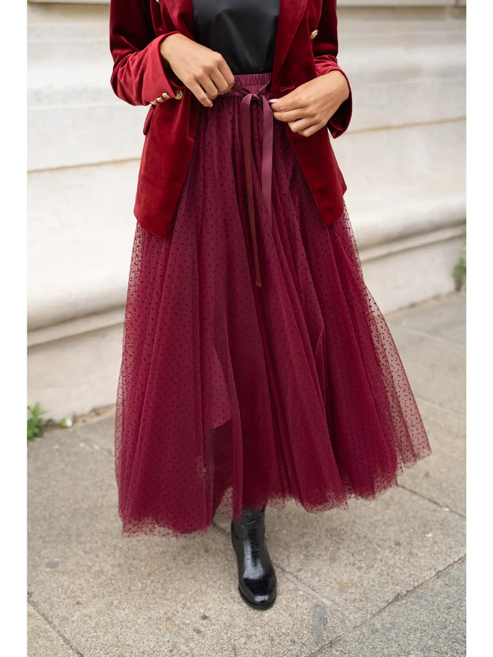 Person wearing a red coat and burgundy tulle skirt on a sidewalk.