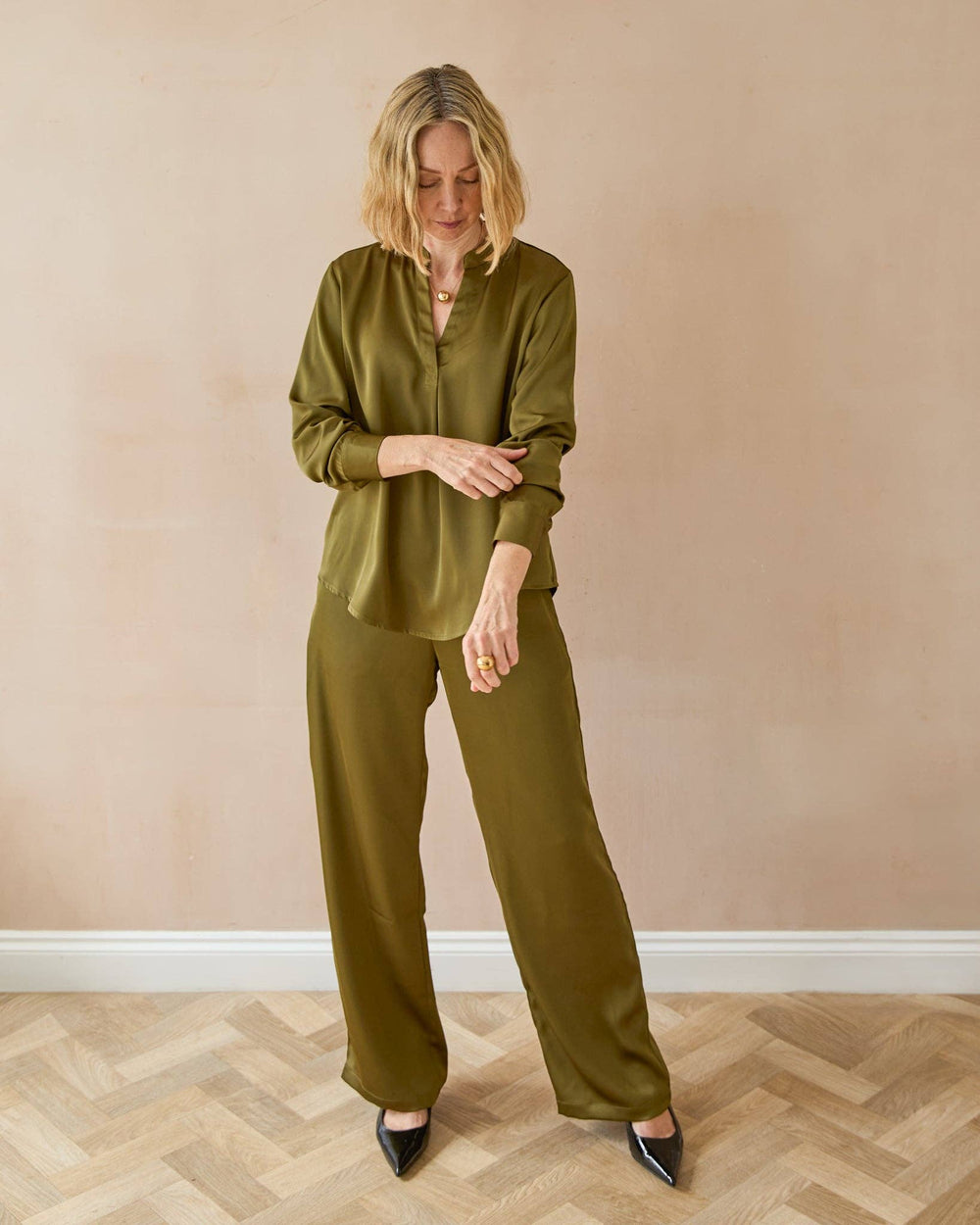Woman wearing an olive green outfit standing against a beige wall.