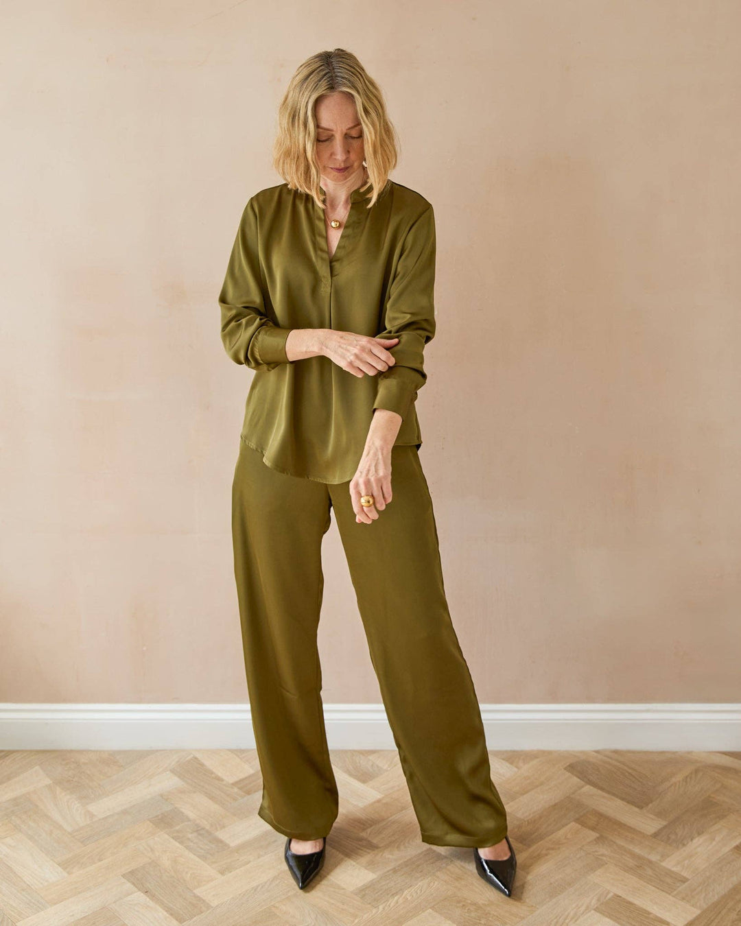 Woman wearing an olive green outfit standing against a beige wall.