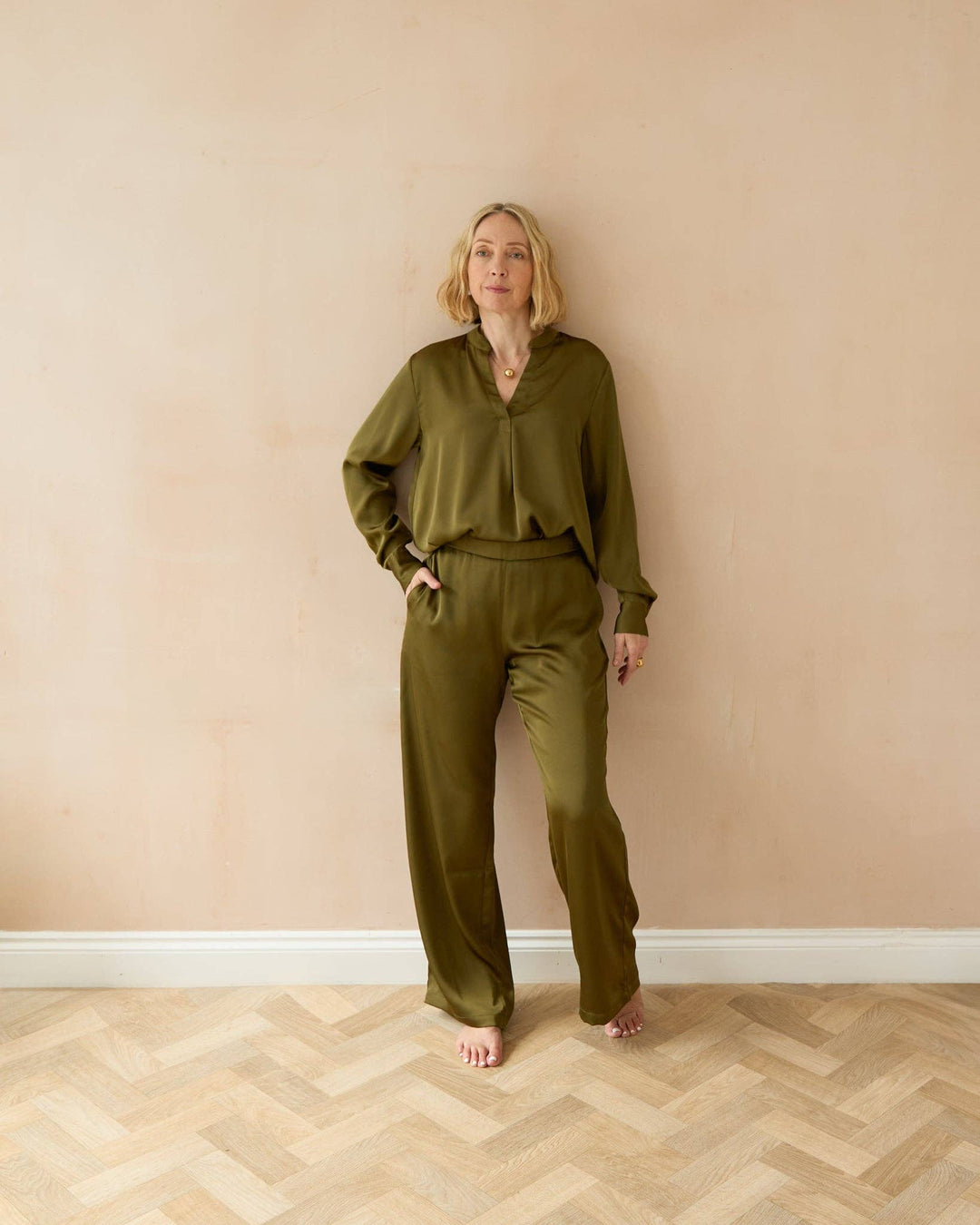 Woman wearing an olive green outfit standing against a beige wall.