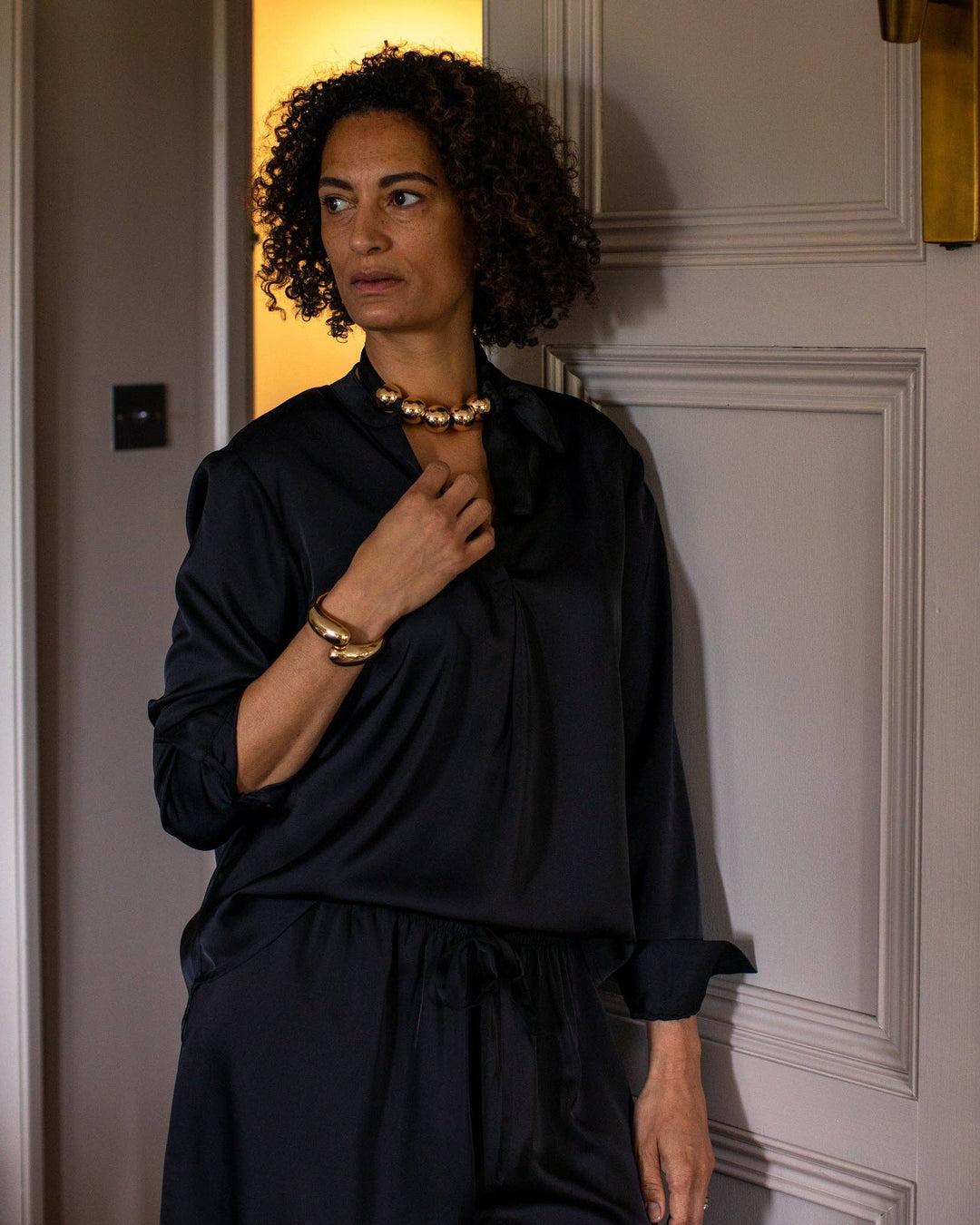 Woman wearing a black outfit with gold jewelry in a room with a door.