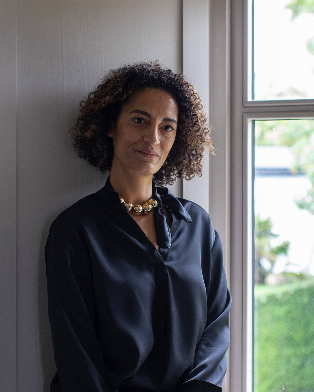 Woman wearing a dark blue shirt with a gold necklace, standing indoors near a window.