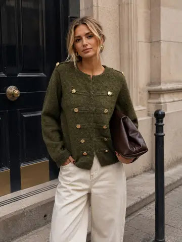 A woman stands on a city street wearing an olive green textured double-breasted jacket with gold buttons, cream wide-leg trousers, and carrying a dark brown leather shoulder bag.