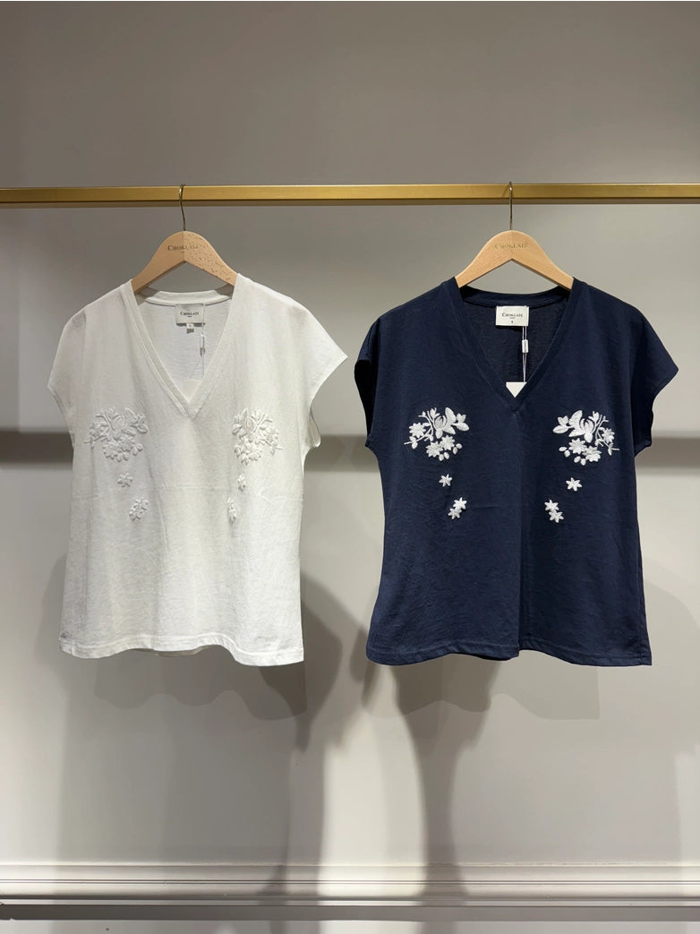 Two shirts, one white and one navy, with floral embroidery on hangers against a neutral background.
