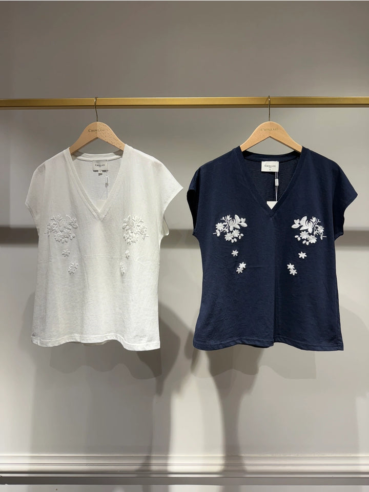 Two shirts, one white and one navy, with floral embroidery on hangers against a neutral background.