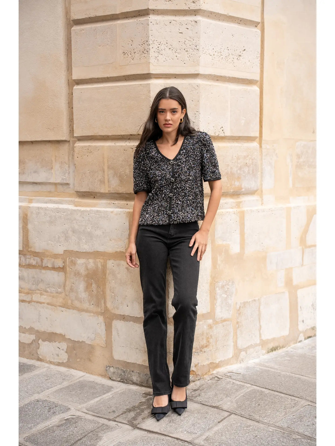 Woman standing against a stone wall wearing a black and white patterned top and black pants.