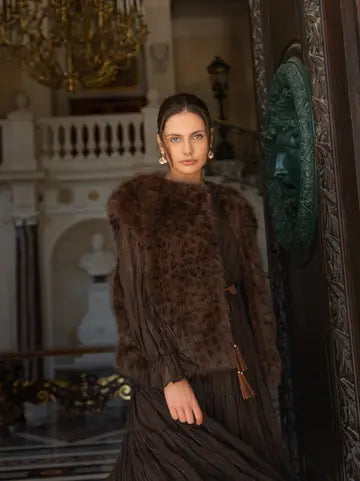 A woman models a luxurious dark brown fur jacket with a leopard-like pattern, worn over a matching dark brown pleated dress in an elegant, historic interior with a chandelier and ornate door.