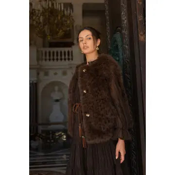 A woman wearing a dark brown fur jacket with gold buttons and a tassel tie, standing in an ornate indoor setting with a staircase and chandelier in the background.