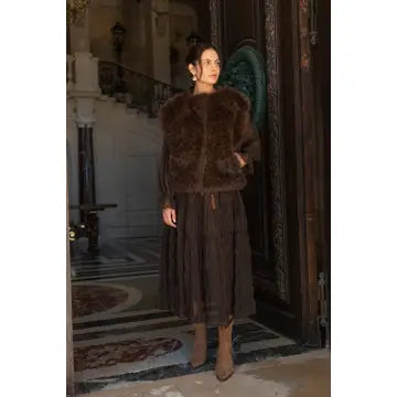 A woman wearing a dark brown fur jacket and a long dark brown pleated skirt stands in a grand, historic interior doorway with ornate wood doors and a staircase in the background.