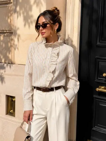 A woman wearing a cream and beige vertically striped blouse with ruffle details, paired with cream trousers and sunglasses, standing outdoors next to a black door.
