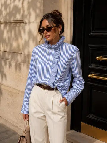 A woman wearing a blue and white vertically striped long-sleeve blouse with a ruffled high neck, styled with cream trousers, a brown belt, and sunglasses, standing outdoors next to a black door.