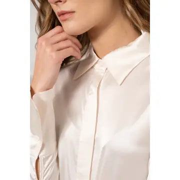 A close-up of a woman wearing a light cream or off-white satin button-up shirt. The image is cropped to show her chin, hand, neck, and the collar and chest area of the shirt, set against a plain light gray background.