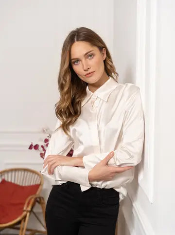 A woman with wavy light brown hair wearing a white satin long-sleeve blouse with a tie-neck detail and black pants, leaning against a white wall in an indoor setting.