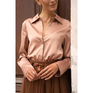 A woman wearing a satin-like long-sleeve button-up blouse in a dusty blush pink color, paired with a brown pleated skirt, standing against a rustic wooden and plaster wall.