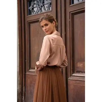 A woman wearing a light beige long-sleeve blouse and a brown pleated skirt, standing in front of a dark brown wooden door.