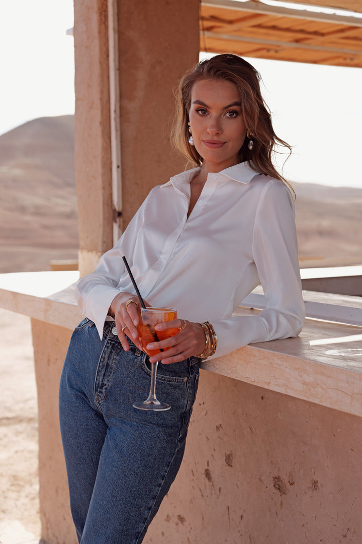 A woman wearing a white long-sleeve button-up shirt and blue jeans leans on a concrete ledge outdoors, holding a cocktail glass. The background features desert hills and warm architectural elements.